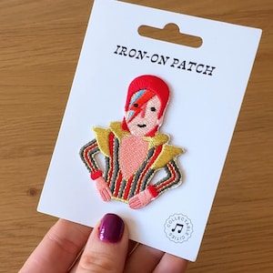Puede incluir: Un parche termoadhesivo bordado que representa una figura con cabello rojo y un rayo en la cara. El parche tiene una combinación de colores dorados, rojos y negros, presentado en una tarjeta blanca con el texto "IRON-ON PATCH".