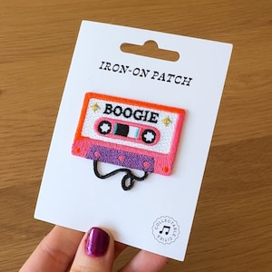Puede incluir: Un parche termoadhesivo bordado con un diseño de casete retro. El parche está bordado con hilo rosa, naranja, morado y blanco, con la palabra "BOOGIE". El parche está adherido a una tarjeta blanca con el texto "IRON-ON PATCH".
