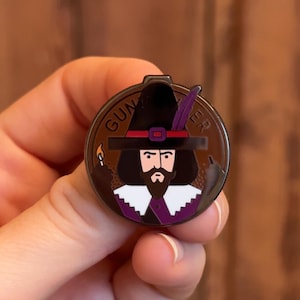 May include: A brown and black enamel pin featuring a cartoon illustration of a man wearing a black hat with a purple feather, a red and white band, and a white collar. The man has a black beard and mustache. The pin says "GUNPOWDER" around the edge.