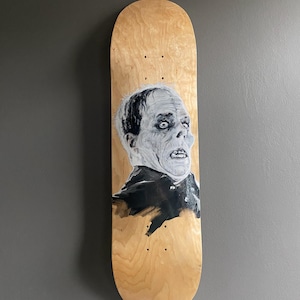 Könnte beinhalten: Ein Skateboard-Deck aus Holz mit einem Schwarzweißbild eines Männergesichts. Der Mann hat einen blassen Teint und einen ernsten Ausdruck. Das Bild ist in einem realistischen Stil gemalt.