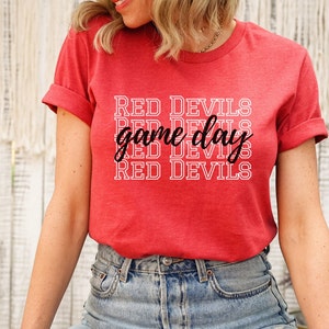 May include: Red t-shirt with rolled-up sleeves. The shirt features the text "RED DEVILS" stacked vertically, with "game day" in a cursive font in the center. The shirt is made of a soft, red fabric.