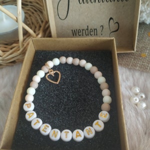 May include: A white beaded bracelet with the word "PATENTE" spelled out in gold letters. The bracelet has a small gold heart charm and is displayed in a brown box.