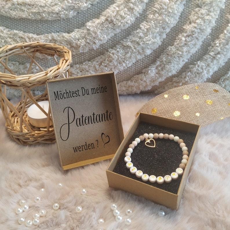 May include: A small gift box with a brown lid and a white interior. The box contains a wooden bead bracelet with a gold heart charm. The box is sitting on a white fuzzy surface. A card with the text "M&ouml;chtest Du meine Patenante werden?" is visible in the background.