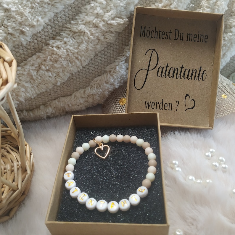 May include: A small gift box with a brown lid and a white interior. The lid has the text "M&ouml;chtest Du meine Patenante werden?" printed on it. The box contains a bracelet with white beads that spell out "Patenante" and a gold heart charm.