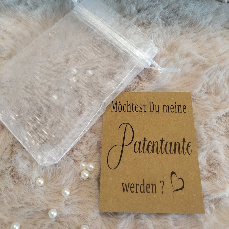 May include: A brown card with the text "M&ouml;chtest Du meine Patenante werden?" with a heart symbol. The card is on a white fluffy surface with a small white organza bag filled with white pearls.