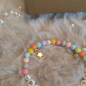May include: A colorful beaded bracelet with a gold star charm. The beads are pastel shades of pink, blue, yellow, and green. The bracelet has white beads with letters spelling out a word.