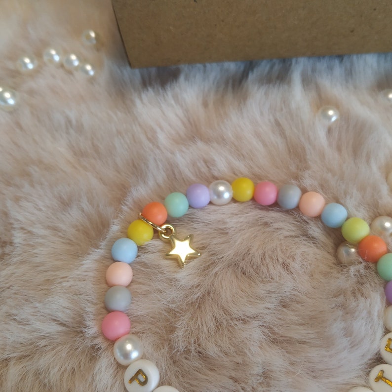 May include: A colorful beaded bracelet with a gold star charm. The beads are pastel shades of pink, blue, yellow, and green. The bracelet has white beads with letters spelling out a word.