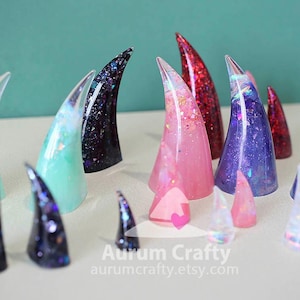 Könnte beinhalten: Eine Sammlung konischer Ringhalter aus Kunstharz in verschiedenen Größen und Farben, darunter Minzgrün, Schwarz mit Glitzer, Pink mit irisierenden Flocken und tiefes Violett mit Funkeln. Jedes Stück hat eine glatte, kegelförmige Form.