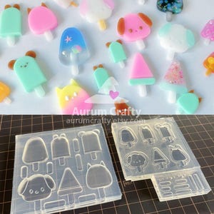 Może przedstawiać: Kolekcja kolorowych zawieszek w kształcie lodów na patyku z żywicy w różnych kształtach, w tym niedźwiedzi, psów i geometrycznych wzorów. Na zdjęciu widoczne są również silikonowe formy z pasującymi kształtami lodów. Widoczny jest tekst "Aurum Crafty aurumcrafty.etsy.com".