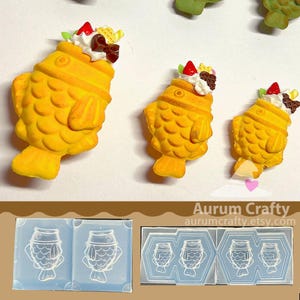 May include: Three yellow, fish-shaped resin charms, each topped with white frosting, a red strawberry, and chocolate accents. The charms are arranged on a white surface. Below are two clear molds with fish-shaped cavities.