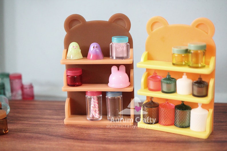 May include: Two miniature wooden shelves with bear ears, one brown and one orange, are filled with miniature jars, bottles, and figurines. The shelves are on a wooden surface.