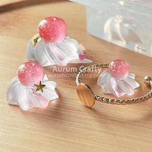 May include: Close-up of three decorative resin pieces. Each piece features a pink, spherical bead atop a white, ruffled base with pink accents. One piece has a gold star, and another is part of a gold bracelet.