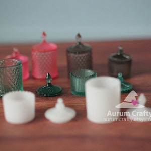 May include: A collection of miniature glass jars with lids in various colours, including green, pink, and white. The jars are arranged on a wooden surface.