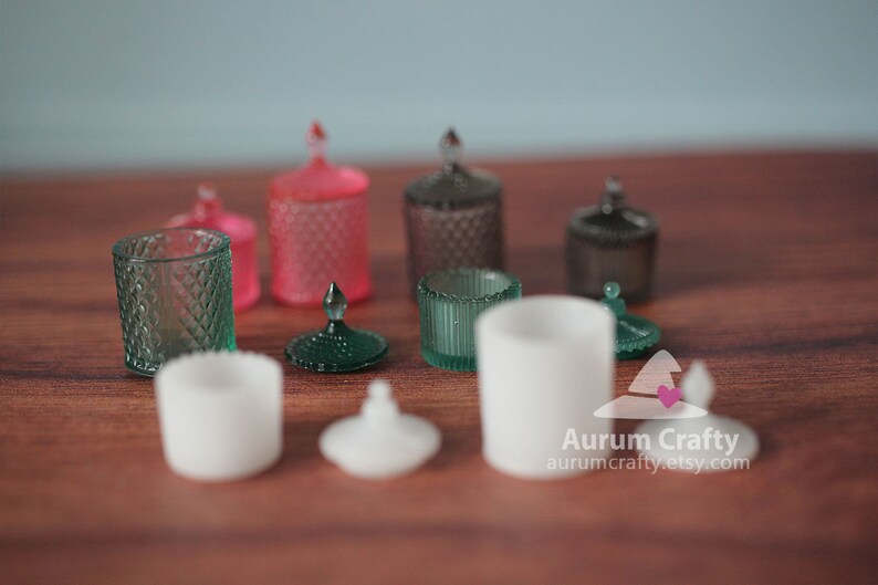 May include: A collection of miniature glass jars with lids in various colours, including green, pink, and white. The jars are arranged on a wooden surface.