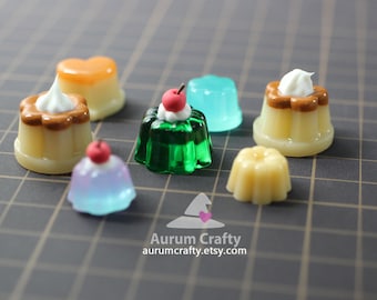Può includere: Assortimento di ciondoli per dessert in miniatura, tra cui una gelatina verde con una ciliegia rossa in cima, una gelatina blu, un budino giallo e due budini marroni con glassa bianca.