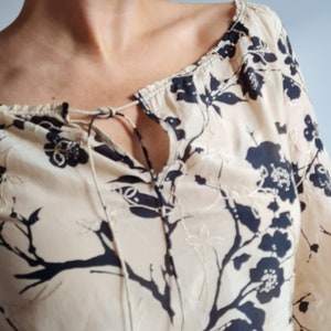 Könnte beinhalten: Eine beige Bluse mit einem Blumendruck mit schwarzen Blüten und Ästen. Die Bluse hat einen Knoten am Ausschnitt.