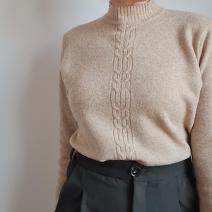 May include: A beige turtleneck sweater with a cable knit design down the front. The sweater is worn with black high-waisted trousers.
