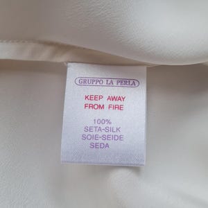 May include: Close-up of a white fabric with a tag. The tag reads "GRUPPO LA PERLA" and "KEEP AWAY FROM FIRE." It also states the fabric is 100% Seta-Silk, Soie-Seide, Seda.