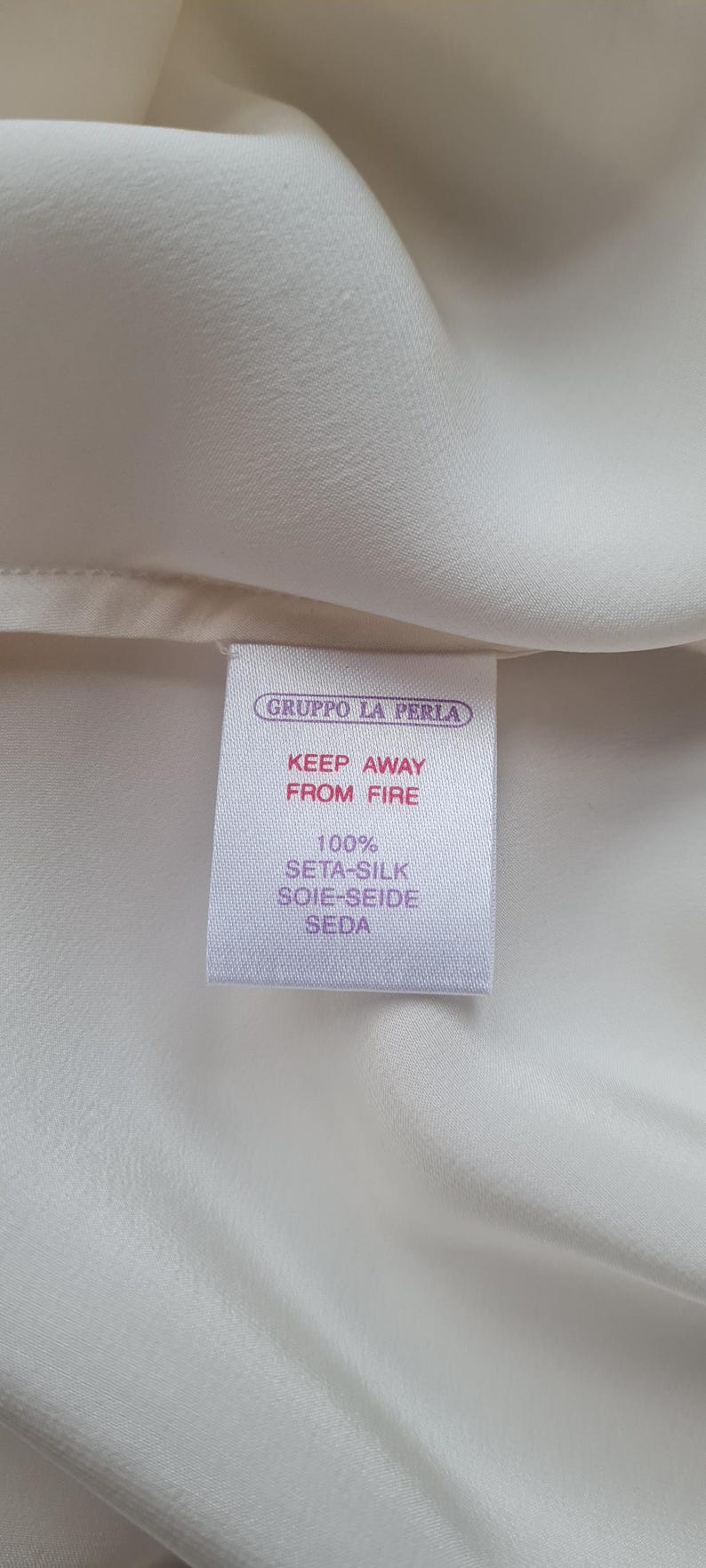 May include: Close-up of a white fabric with a tag. The tag reads "GRUPPO LA PERLA" and "KEEP AWAY FROM FIRE." It also states the fabric is 100% Seta-Silk, Soie-Seide, Seda.