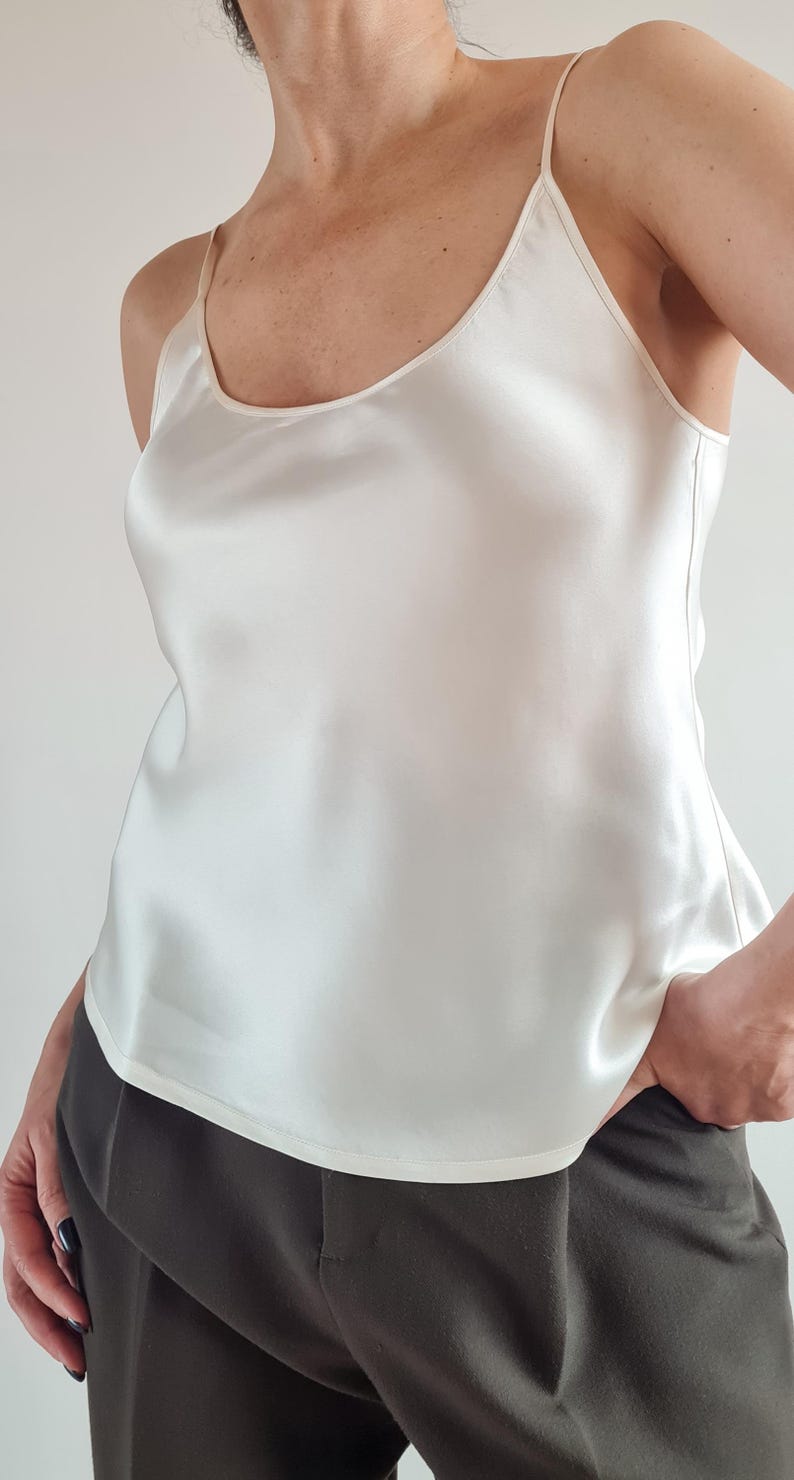 May include: A cream-colored camisole with thin straps. The top is made of a silky material and has a slight sheen. The model is wearing dark green pants. The top is a simple, elegant design.