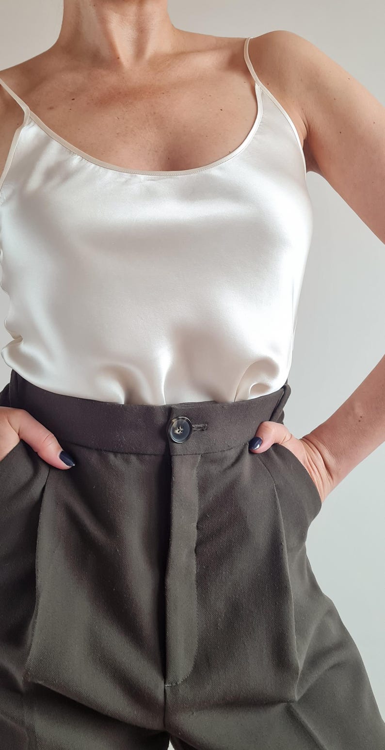 May include: Ivory silk camisole top with spaghetti straps paired with dark olive green high-waisted trousers. The trousers have a button closure and a tailored fit, creating a sophisticated ensemble. The outfit is simple and elegant.