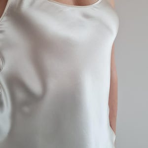 May include: A cream-colored satin camisole with thin spaghetti straps. The top has a scoop neckline and a smooth, lustrous finish. The garment is a solid color and appears to be made of a lightweight fabric.
