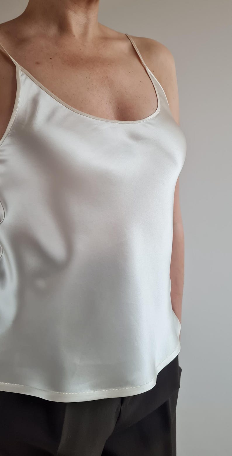 May include: A cream-colored satin camisole with thin spaghetti straps. The top has a scoop neckline and a smooth, lustrous finish. The garment is a solid color and appears to be made of a lightweight fabric.
