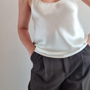 May include: Ivory-colored silk camisole with thin straps and a scoop neckline. The top is paired with dark gray pleated trousers. The outfit is simple and elegant, suitable for various occasions.