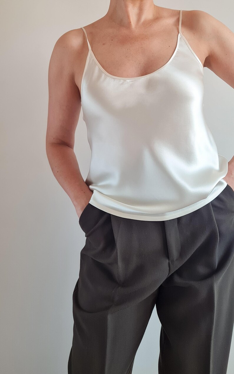May include: Ivory-colored silk camisole with thin straps and a scoop neckline. The top is paired with dark gray pleated trousers. The outfit is simple and elegant, suitable for various occasions.