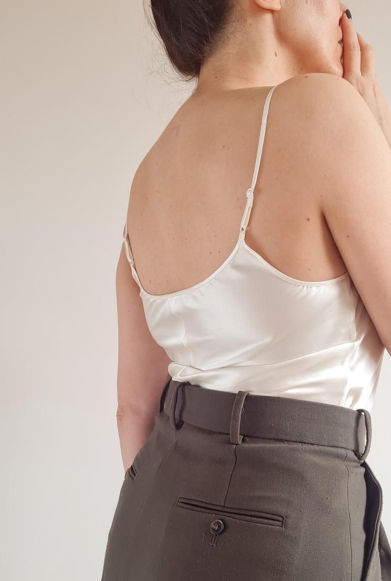 May include: A white satin camisole top with thin spaghetti straps is paired with dark gray high-waisted trousers. The top has a subtle sheen, and the trousers feature a tailored fit with a belt.