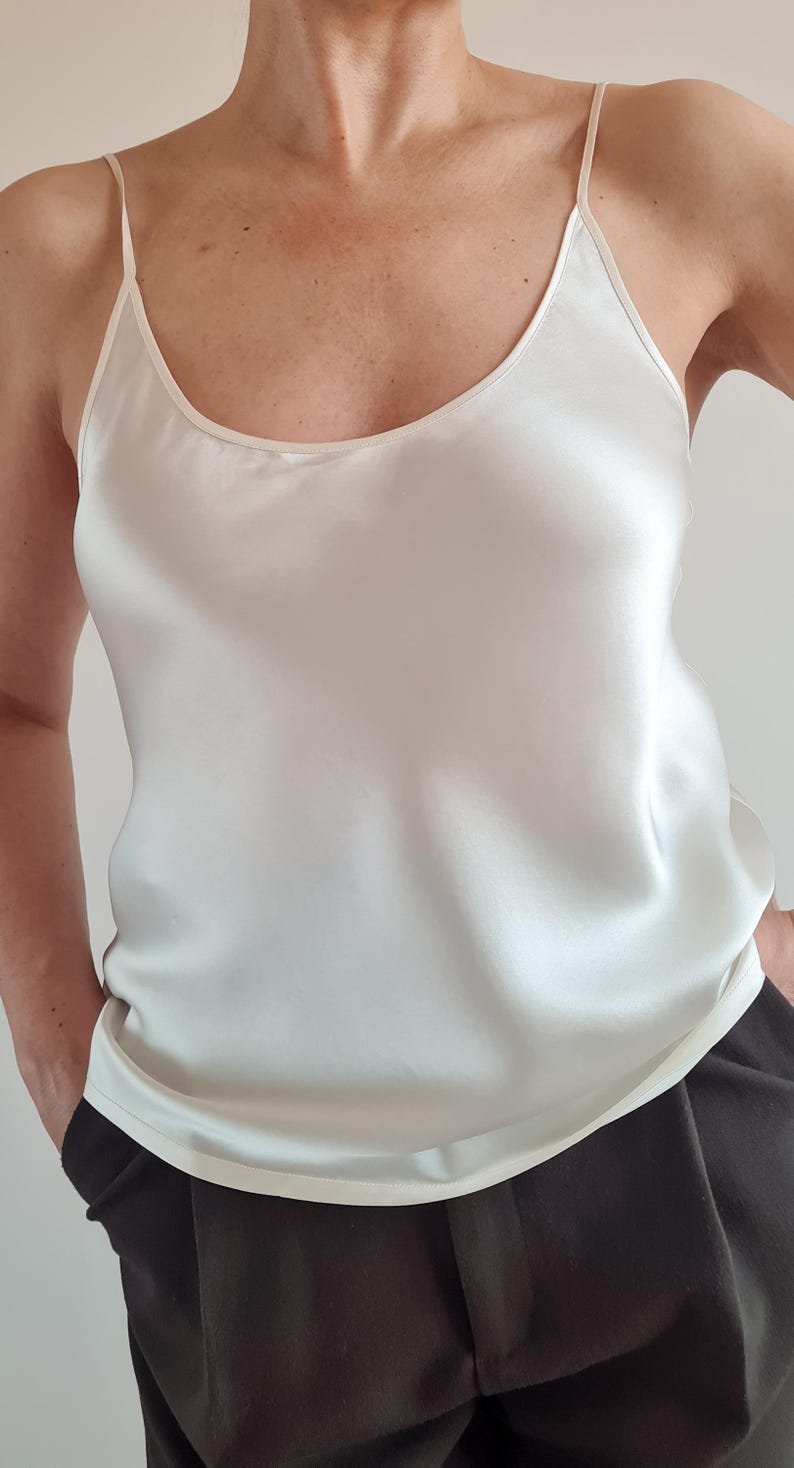 May include: Ivory-colored camisole top with thin spaghetti straps and a scoop neckline. The silky fabric drapes smoothly. The model is wearing black pants. The top is a neutral color, suitable for layering or wearing on its own.