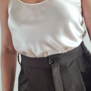 May include: A cream-colored camisole top with spaghetti straps paired with dark brown high-waisted trousers. The top has a silky sheen, and the trousers feature a button closure and a tailored fit. The outfit is simple and elegant.