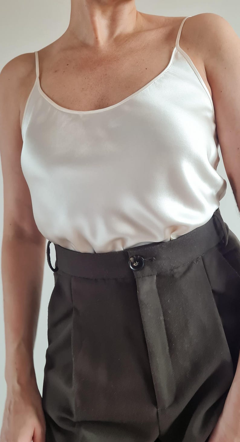 May include: A cream-colored camisole top with spaghetti straps paired with dark brown high-waisted trousers. The top has a silky sheen, and the trousers feature a button closure and a tailored fit. The outfit is simple and elegant.