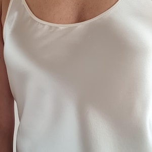 May include: Ivory-colored camisole top with thin spaghetti straps. The top has a smooth, silky texture and a scoop neckline. The fabric drapes softly. The garment is a solid color and appears to be made of a lightweight material.
