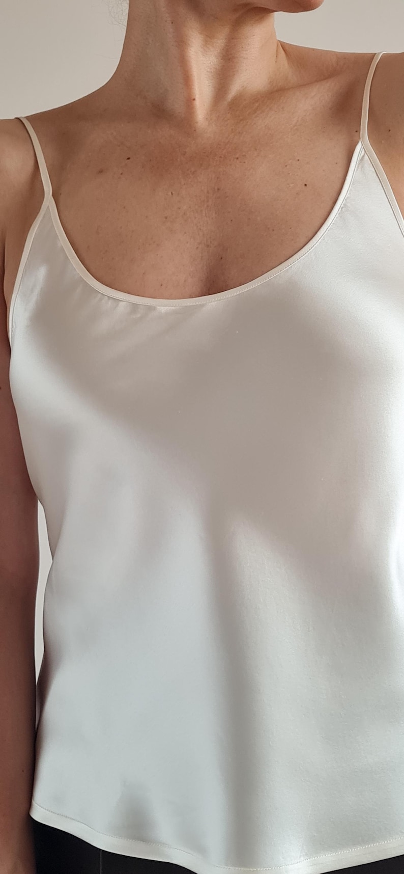 May include: Ivory-colored camisole top with thin spaghetti straps. The top has a smooth, silky texture and a scoop neckline. The fabric drapes softly. The garment is a solid color and appears to be made of a lightweight material.