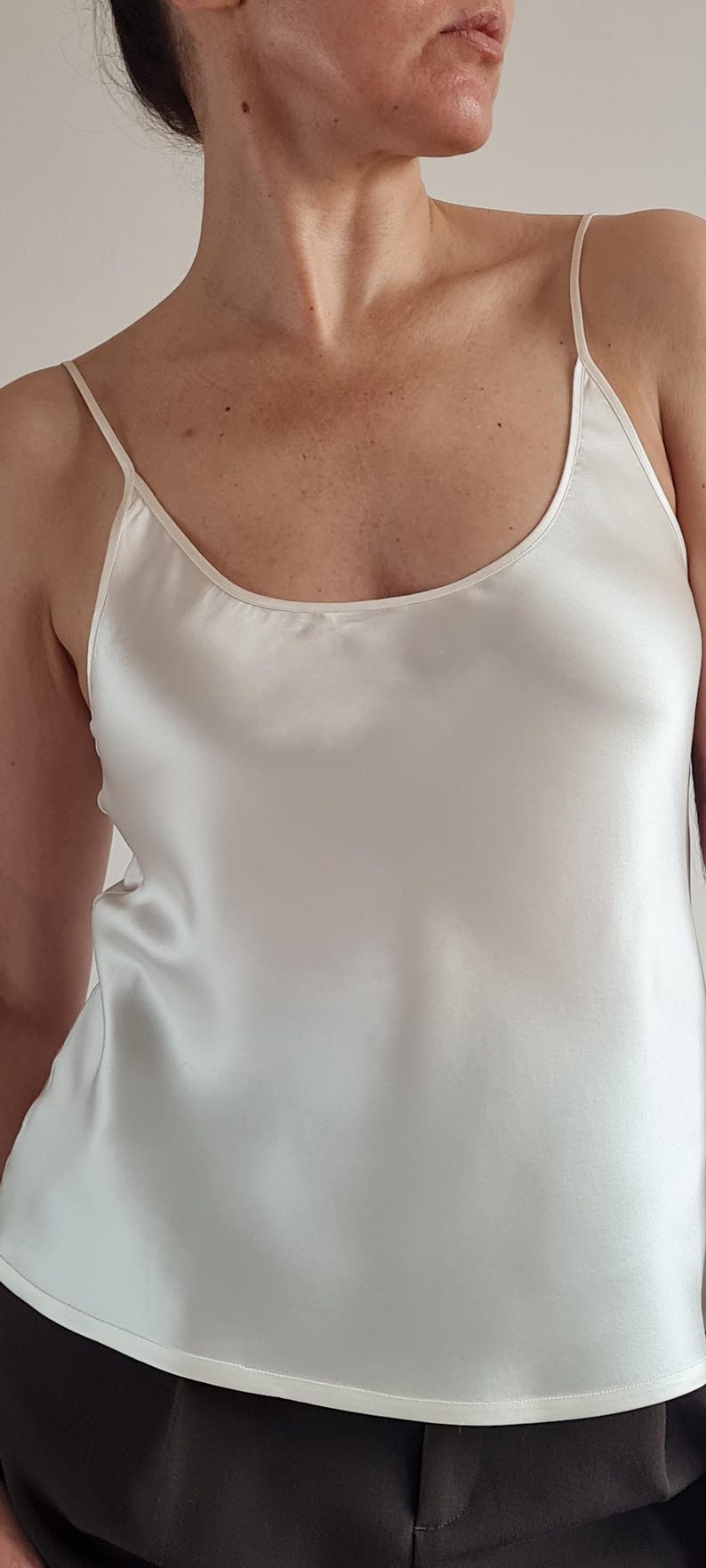 May include: Ivory-colored camisole top with thin spaghetti straps. The silky fabric has a subtle sheen and drapes smoothly. The top has a scoop neckline and is a neutral color, suitable for layering or wearing alone.
