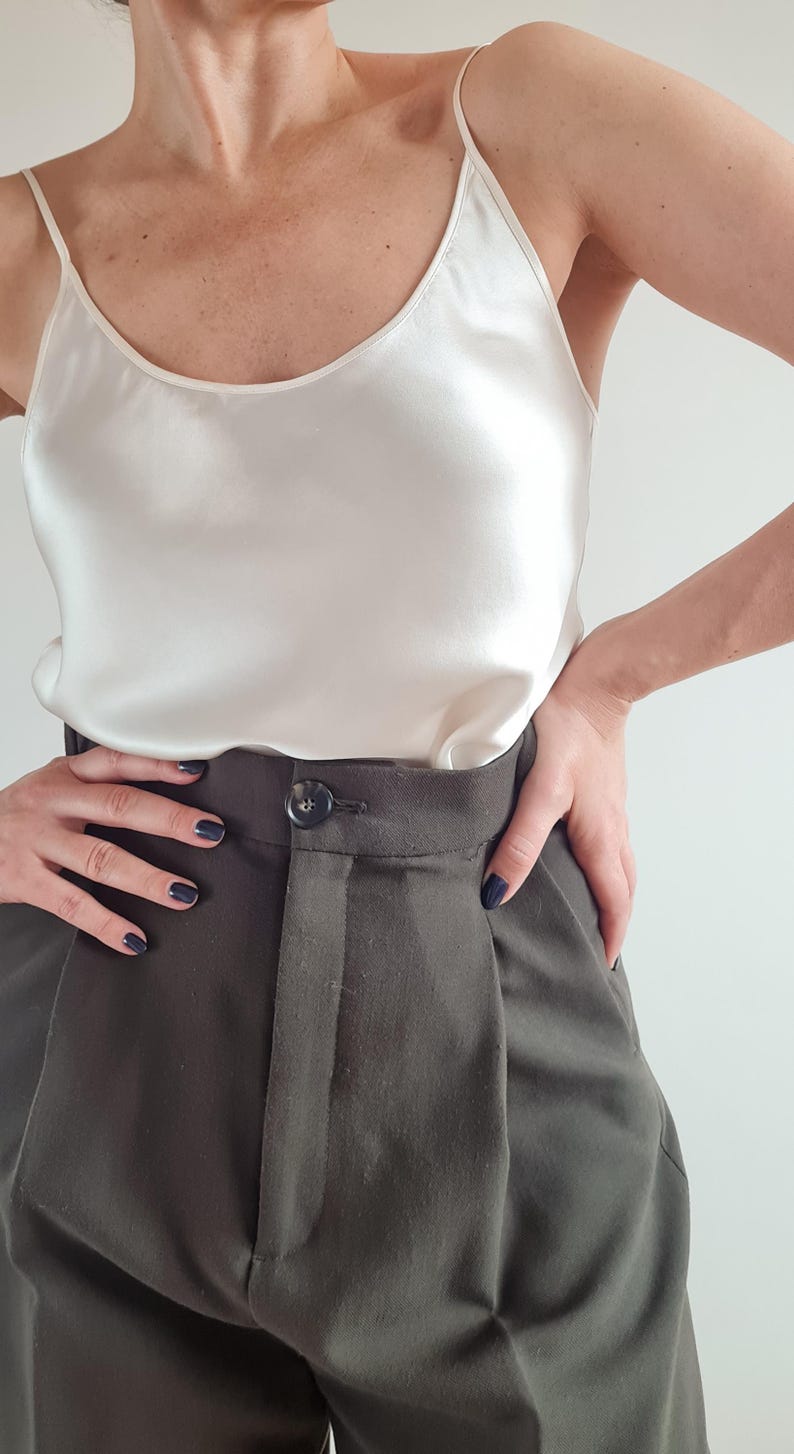 May include: A cream-colored camisole top with spaghetti straps paired with dark gray trousers. The trousers have a button closure and pleats. The outfit is simple and elegant.