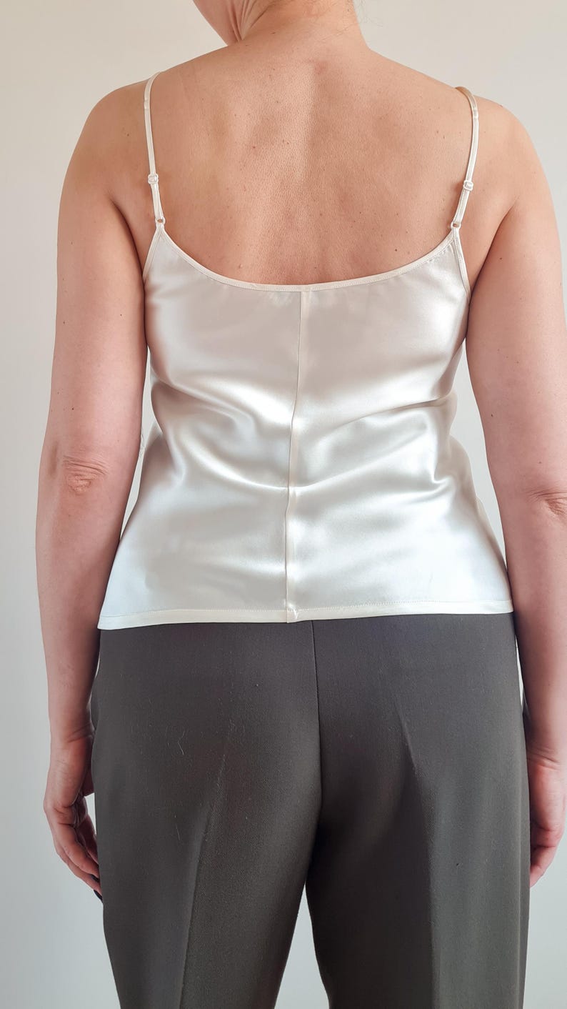 May include: Ivory-colored camisole top with thin spaghetti straps. The silky fabric has a subtle sheen and a simple, elegant design. The top is paired with dark gray trousers, creating a classic and versatile outfit.