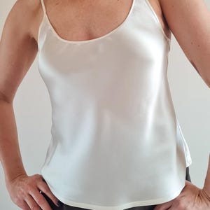 May include: Ivory-colored camisole top with thin spaghetti straps and a scoop neckline. The silky fabric drapes smoothly. The top is a solid color and has a simple, elegant design. The garment is a versatile piece for layering or wearing on its own.