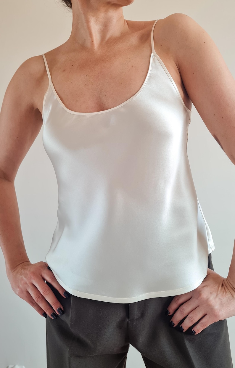 May include: Ivory-colored camisole top with thin spaghetti straps and a scoop neckline. The silky fabric drapes smoothly. The top is a solid color and has a simple, elegant design. The garment is a versatile piece for layering or wearing on its own.