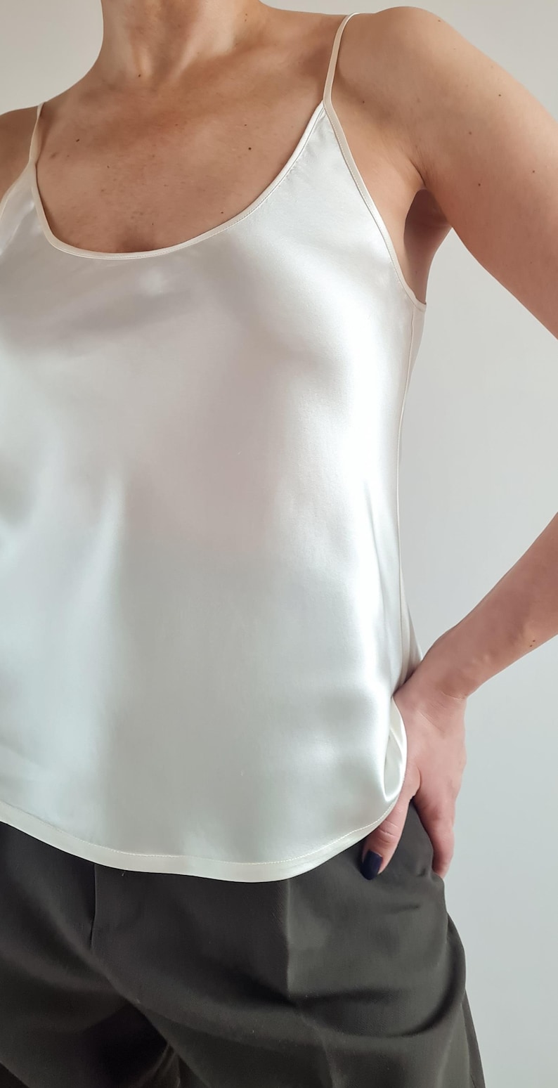 May include: A white satin camisole with thin spaghetti straps. The top has a scoop neckline and a smooth, shiny texture. The camisole is paired with dark green trousers.