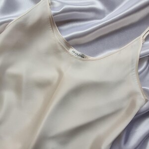 May include: A cream-colored silk camisole with thin straps, resting on a backdrop of draped, silvery-gray fabric. The camisole has a scoop neckline and the brand name "MALIZIA" is visible on a small tag.