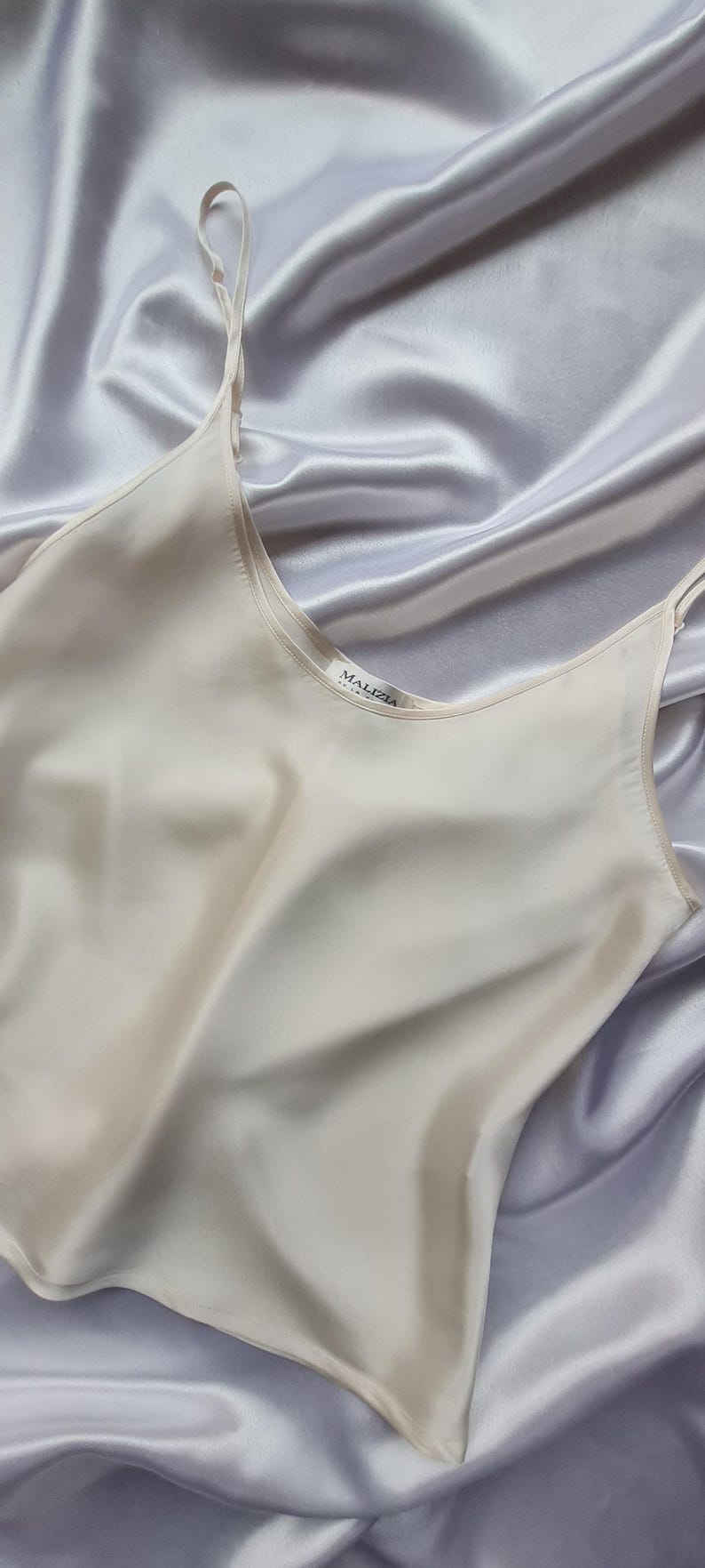 May include: A cream-colored silk camisole with thin straps, resting on a backdrop of draped, silvery-gray fabric. The camisole has a scoop neckline and the brand name "MALIZIA" is visible on a small tag.