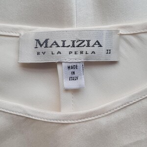 May include: Cream-colored garment with a label that reads "MALIZIA BY LA PERLA" and "MADE IN ITALY." The label is rectangular with dark gray text. The garment appears to be made of a smooth, silky fabric.
