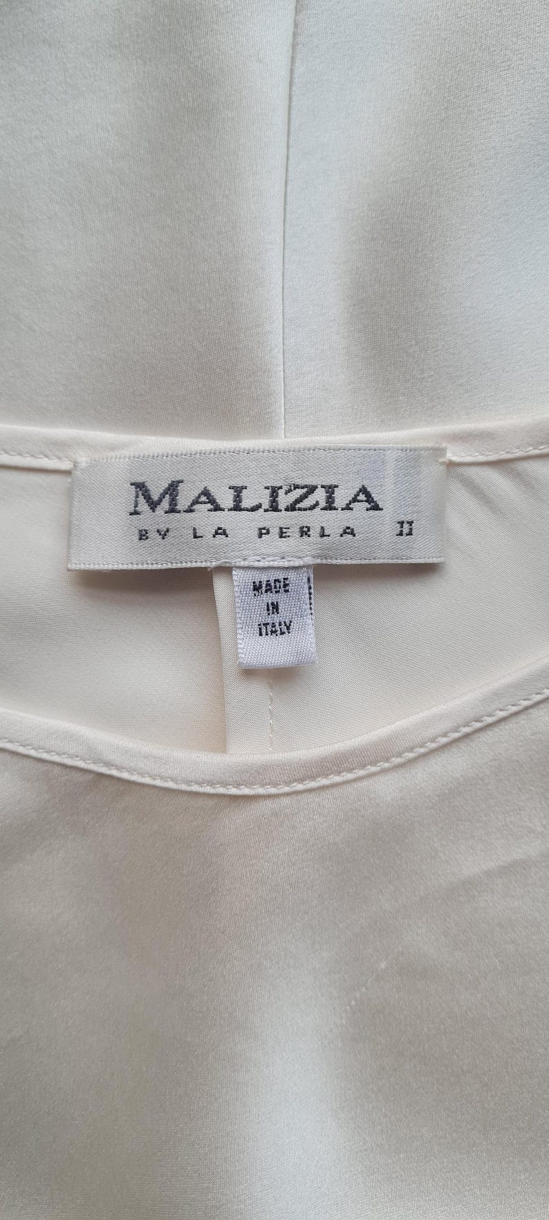 May include: Cream-colored garment with a label that reads "MALIZIA BY LA PERLA" and "MADE IN ITALY." The label is rectangular with dark gray text. The garment appears to be made of a smooth, silky fabric.