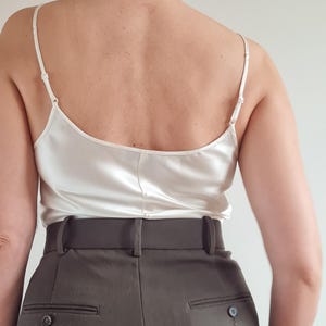 May include: A cream-colored silk camisole with thin spaghetti straps. The camisole is paired with high-waisted, dark olive-green trousers. The trousers have a tailored fit with a buttoned back pocket.
