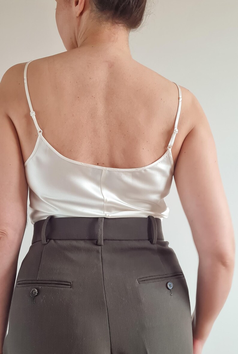 May include: A cream-colored silk camisole with thin spaghetti straps. The camisole is paired with high-waisted, dark olive-green trousers. The trousers have a tailored fit with a buttoned back pocket.