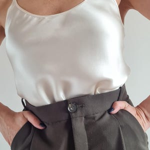May include: A cream-colored silk camisole top with spaghetti straps paired with dark brown high-waisted trousers. The trousers have a button closure and pockets. The outfit is simple and elegant.