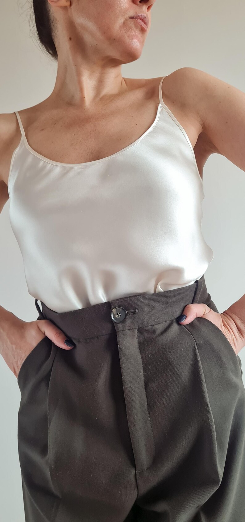 May include: A cream-colored silk camisole top with spaghetti straps paired with dark brown high-waisted trousers. The trousers have a button closure and pockets. The outfit is simple and elegant.
