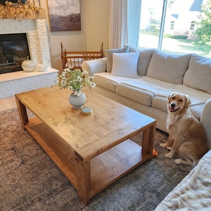 Puede incluir: Un salón con una gran mesa de centro rectangular de madera. La mesa está sobre una alfombra estampada, con un sofá beige y un perro golden retriever. Una chimenea y obras de arte son visibles en el fondo. La mesa mide aproximadamente 120 cm de largo.